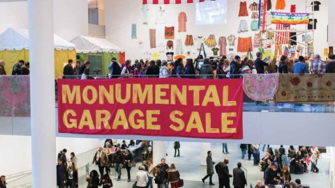 The Monumental Garage Sale at the Museum of Modern Art with a line of people mulling around and several carpets displayed over a glass railing.