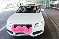 A white car with a pink mustache attached to it's front grill with two people riding.