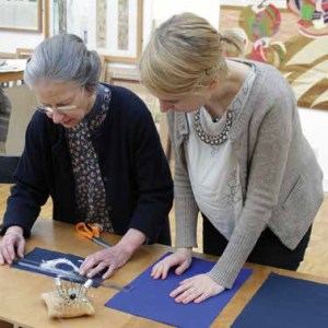 Two women stand at a ttable measuring fabric with a set of pins and other sewing tools around.