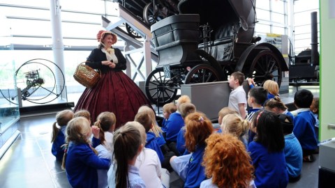 An image of children seated on the floor in a museum listening to a speaker in period dress. The speaker is wearing a bonnet, dress, and carrying a basket.