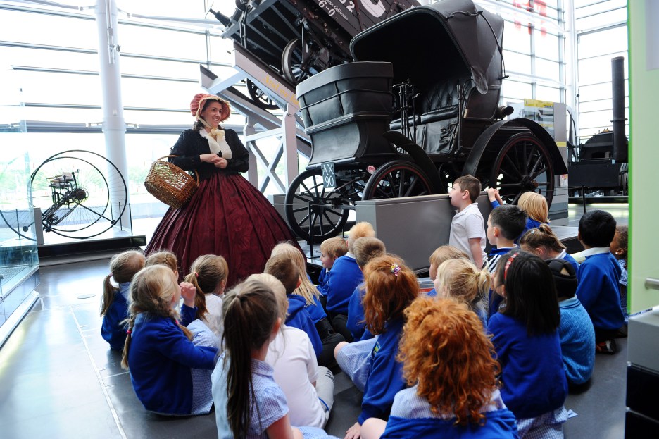 An image of children seated on the floor in a museum listening to a speaker in period dress. The speaker is wearing a bonnet, dress, and carrying a basket.