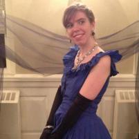 Picture of author Alli Hartley in a blue dress with long black gloves and fancy jewelry, standing in front of a black tulle fabric.