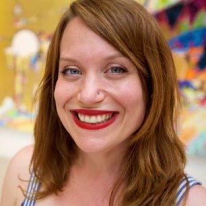 Closeup picture of Mara Kurlandsky smiling with red lipstick, blues eyes, red hair in front of colorful background