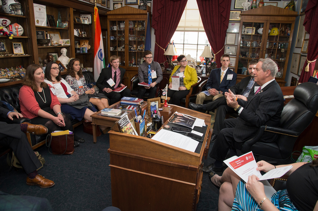 Nine people gather around a wooden desk wearing suits and listening to a representative talk.