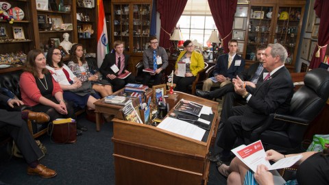 Nine people gather around a wooden desk wearing suits and listening to a representative talk.
