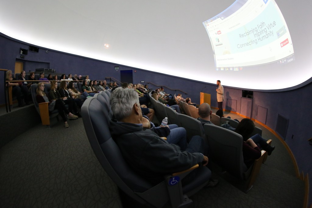 Image of a large group watching a full screen presentation with a man speaking.