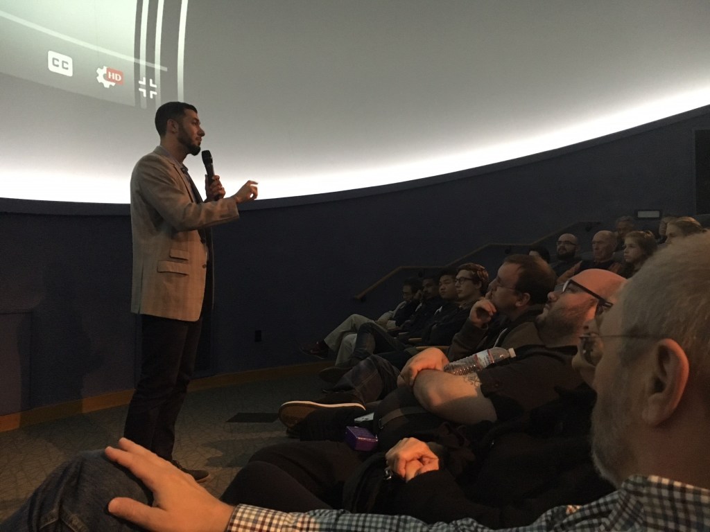 Image of a man standing before a group of interested attendees watching a screen.