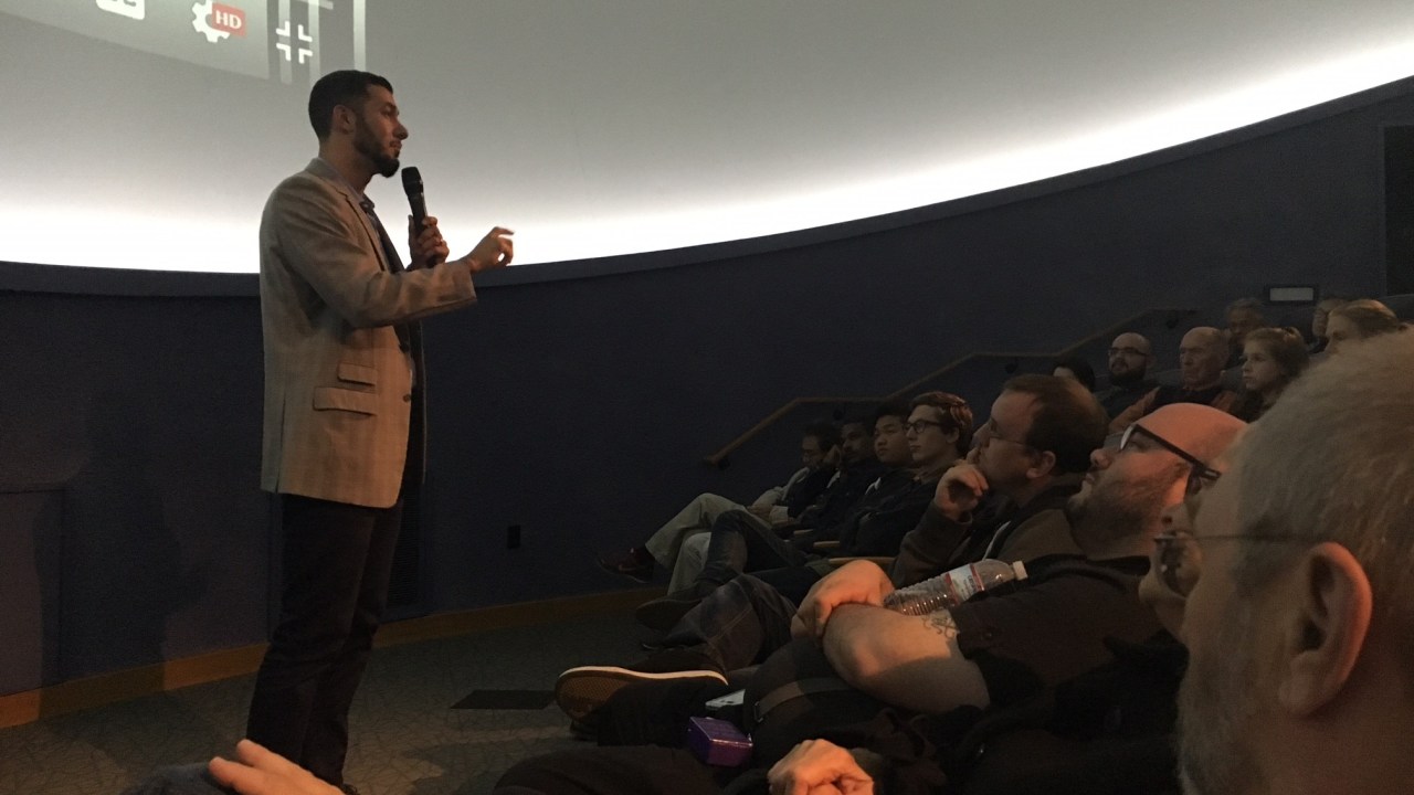 Image of a man standing before a group of interested attendees watching a screen.