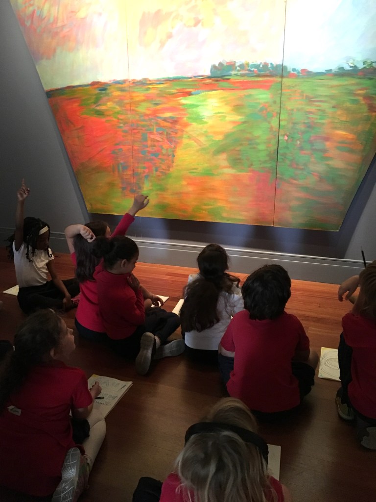 Several students sit cross-legged looking at a painting on the wall.