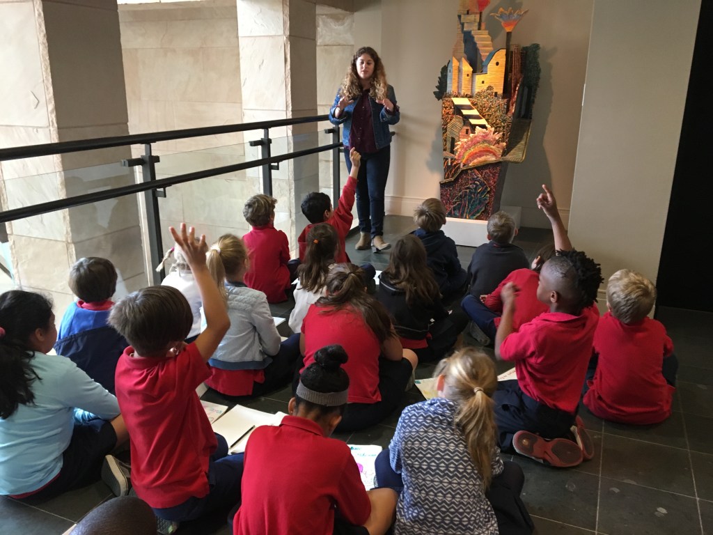 A teach stands discussing a sculpture with a group of students in front of her.