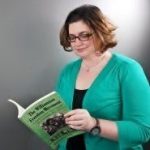 Woman standing in a green sweater reading a textbook wearing glasses with ear length brown hair