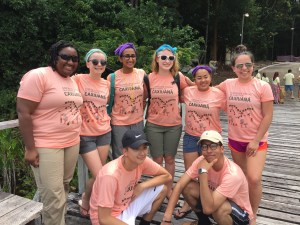 8 people stand and kneel on a wooden walkway in front of trees