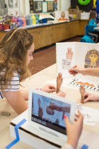 A young girl leans over a table with a miniature mummy sculpture while other children (out of screen) use technology or also hold mummy's. 