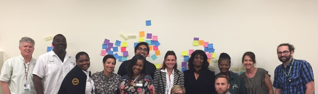 Group of diverse individuarls standing and sitting in front of a white wall with post-it notes on it