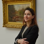 Woman stands smiling with her arms folded in front of her standing in front of a painting in a gilded frame she has long brown hair and is wearing a black blazer and grey shirt