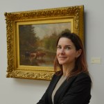 Woman stands smiling with her arms folded in front of her standing in front of a painting in a gilded frame she has long brown hair and is wearing a black blazer and grey shirt