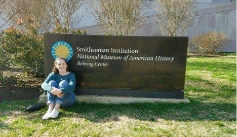 Woman sits with her kness in front of her in ftron of the sign for the Smithsonian Museum of American History
