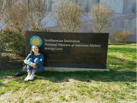 Woman sits with her kness in front of her in ftron of the sign for the Smithsonian Museum of American History
