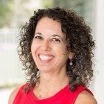 Woman smiling with shoulder length curly brown hair wearing a red sleeveless top