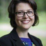 Woman smiling short brown hair wearing glasses a black blazer and blue shirt