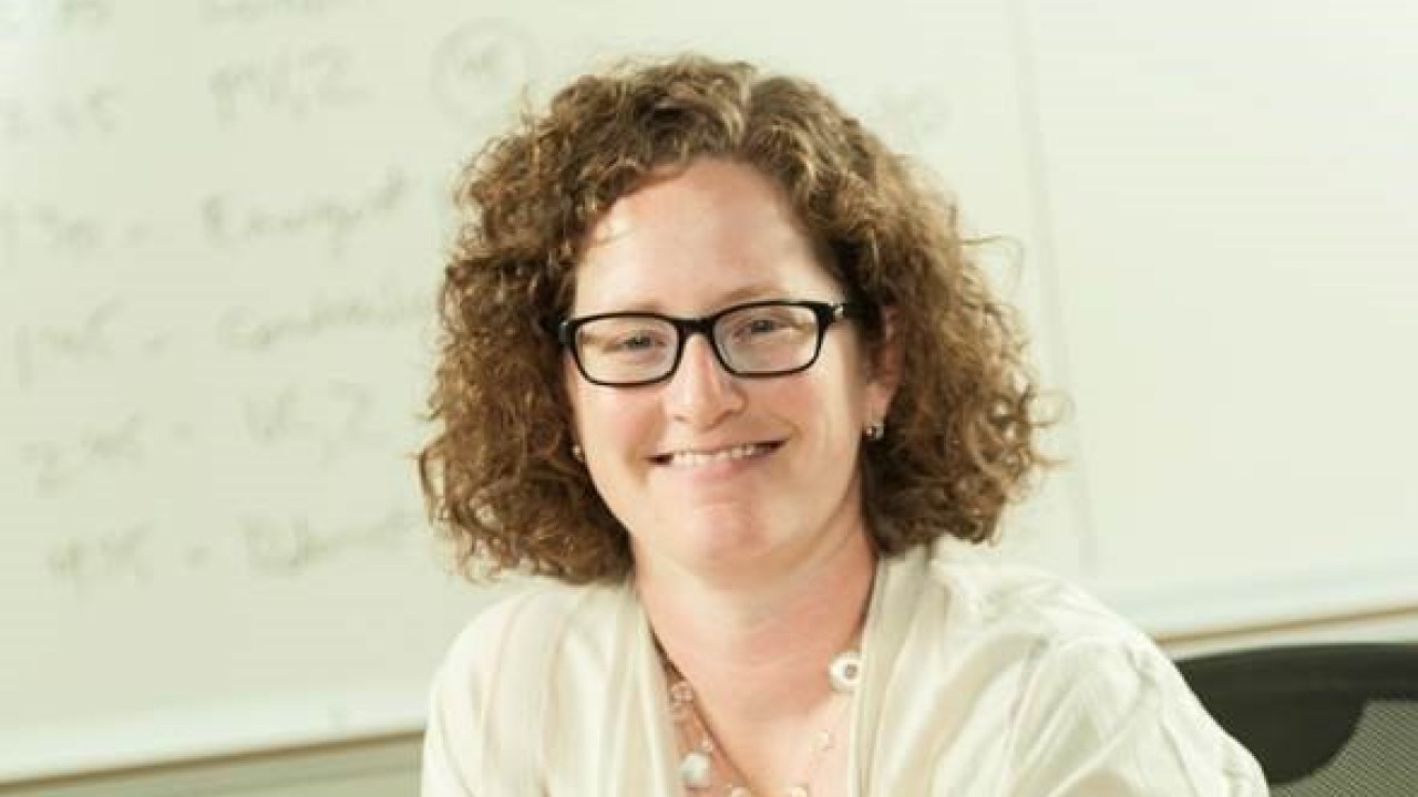 A picture of Sarah Cohn leaning against a desk and smiling at the camera.