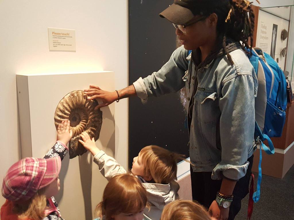 Several young children stand around a mollusk fossil with their hands outstreached. An adult shows them it is okay.