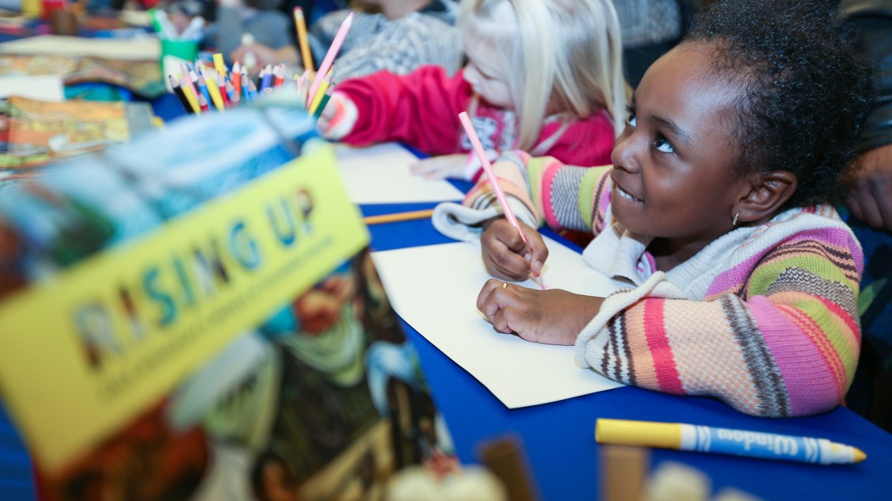 A group of young children sit at a long table coloring and writing. One youngster is smiling up at someone off-camera.