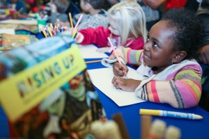 A group of young children sit at a long table coloring and writing. One youngster is smiling up at someone off-camera.