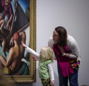 A young child points at part of a person in the painting while an adult stoops intently toward them.