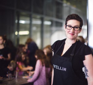 A woman in a blck top with a black apron that says Wellin on it stands smilling a the camera with a group of children in the background. 