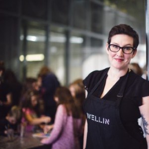 A woman in a blck top with a black apron that says Wellin on it stands smilling a the camera with a group of children in the background.