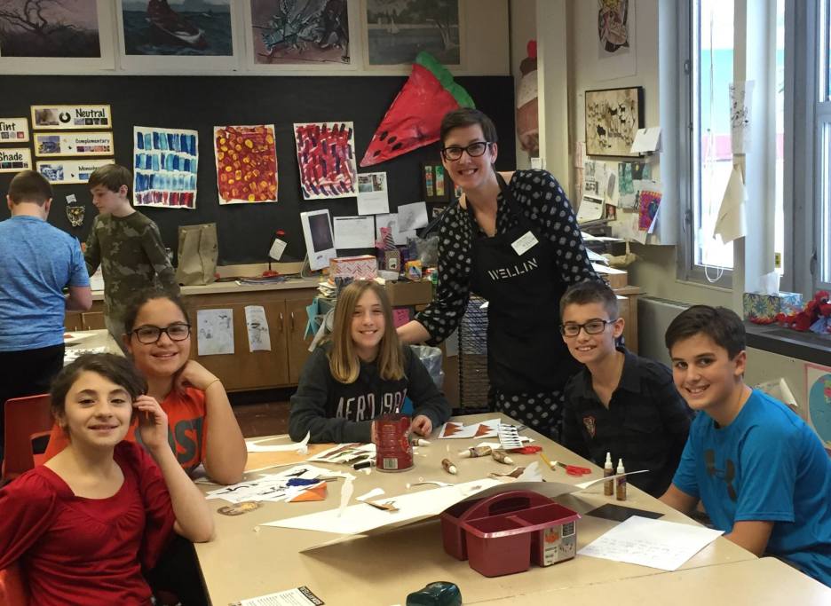 Several young people sit around a table their teacher stands behind them smiling.