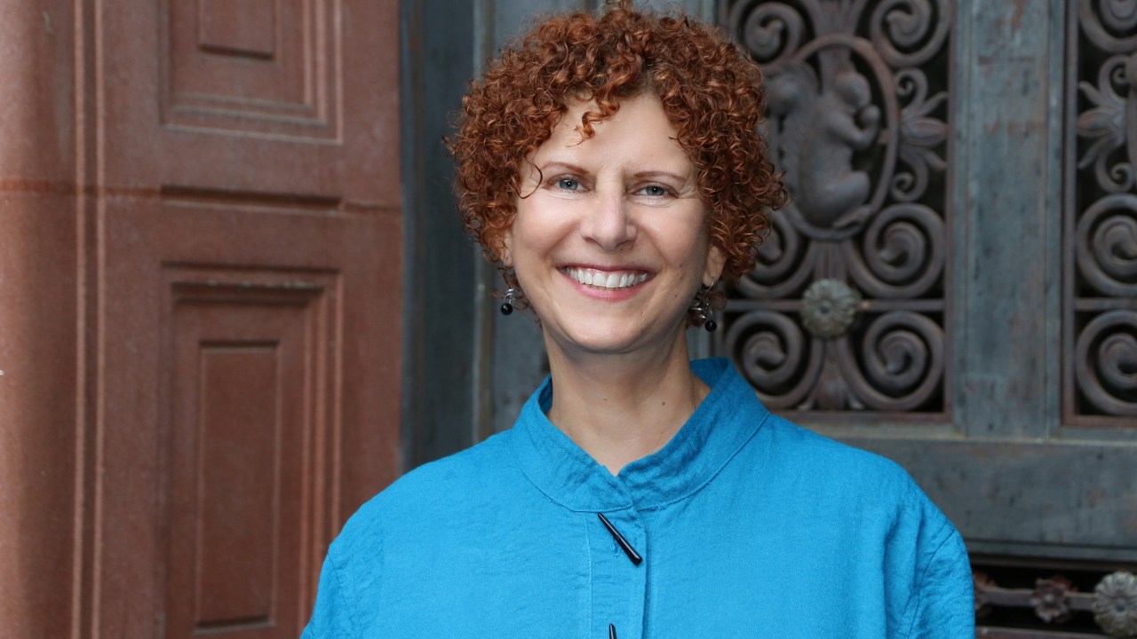 Commissioner Jill Hartz stands in front of an architectural element (perhaps a door) smiling with short red curly hair wearing a blue top