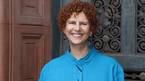 Commissioner Jill Hartz stands in front of an architectural element (perhaps a door) smiling with short red curly hair wearing a blue top
