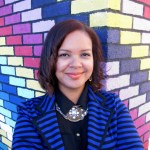 Woman smiling in front of a colorful brick wall wearing a striped blue jacket and black shirt