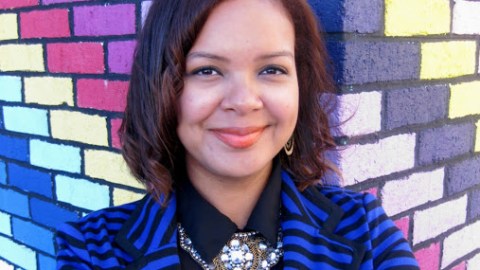 Woman smiling in front of a colorful brick wall wearing a striped blue jacket and black shirt