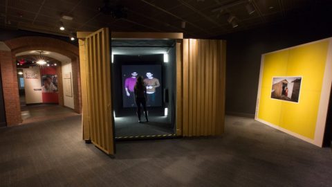 A shipping container painted yellow sits in a gallery with a video screen at the end where two individuals are shown on the screen while another interacts with them in real life.