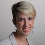 Headshot of a woman smiling with short blond colored hair wearing long earrings and a white blazer