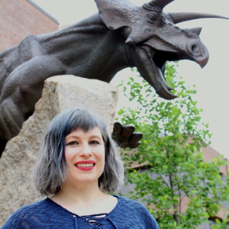 Woman stands in front of a dinosaur statue (possibly bronz) ouside with purple or grey hair wearing a blue shirt