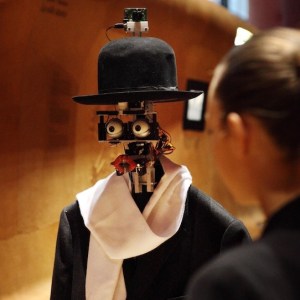 A woman stands speaking with a robot with no face but has eyes an lips wearing a top hat and scarf with a jacket.