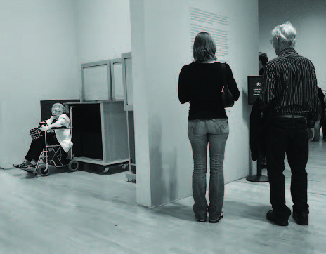 An older woman sits in a wheelchair in what looks like a coat room while two able bodied individuals stand reading wall text in a gallery around the corner