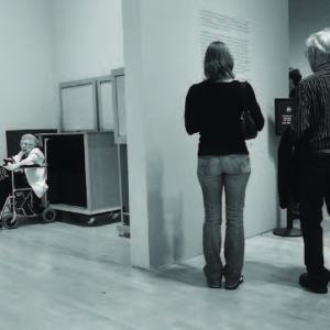 An older woman sits in a wheelchair in what looks like a coat room while two able bodied individuals stand reading wall text in a gallery around the corner