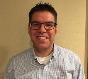Headshot of a man smiling at the camera wearing black rimmed glasses wearing a button down shirt with a white t-shirt underneath.