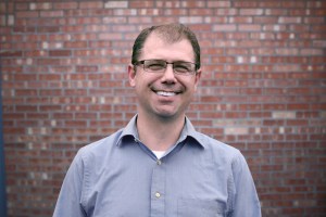 A man stands smiling in front of a brick wall wearing a blue button down shirt