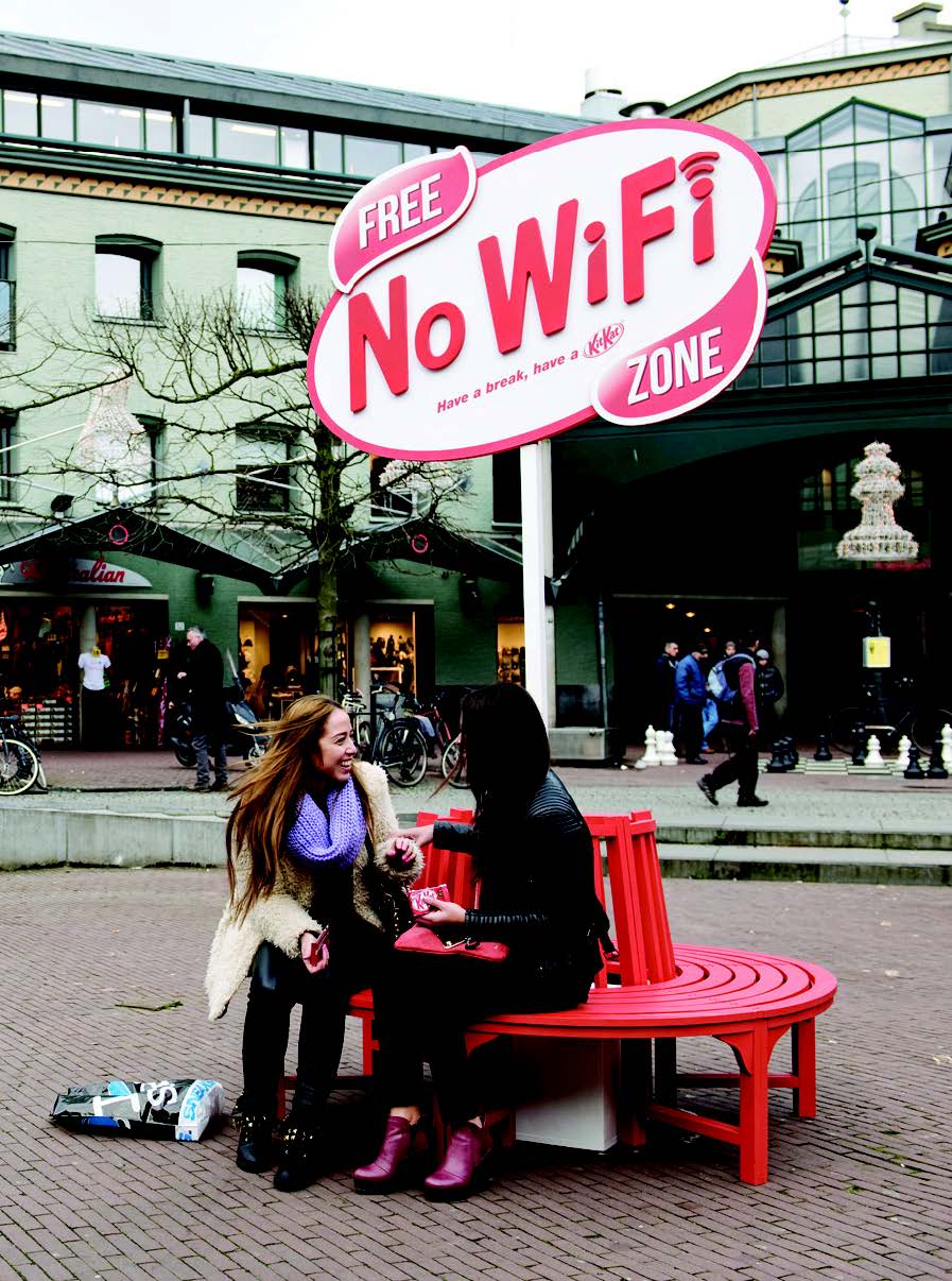 Two women sit on a round bench in front of a Free No WiFi Zone sign