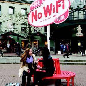 Two women sit on a round bench in front of a Free No WiFi Zone sign