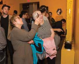 Two women are in the foreground looking at an installation and smiling. One is taking a picture on her phone.