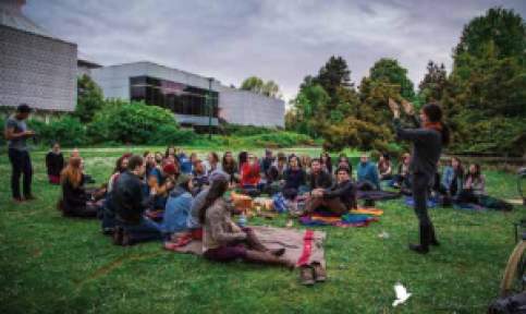 A group of people sit outside on a grassy area an dlook at a person presenting