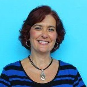 A woman stands smiling in front of a blue wall with short brown hair and a striped blue and black shirt