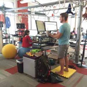 A man stands at a standing desk another walks on a treadmill desk